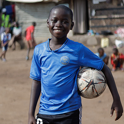 Jordan uit Uganda kijkt lachend in de camera met een bal onder zijn arm.